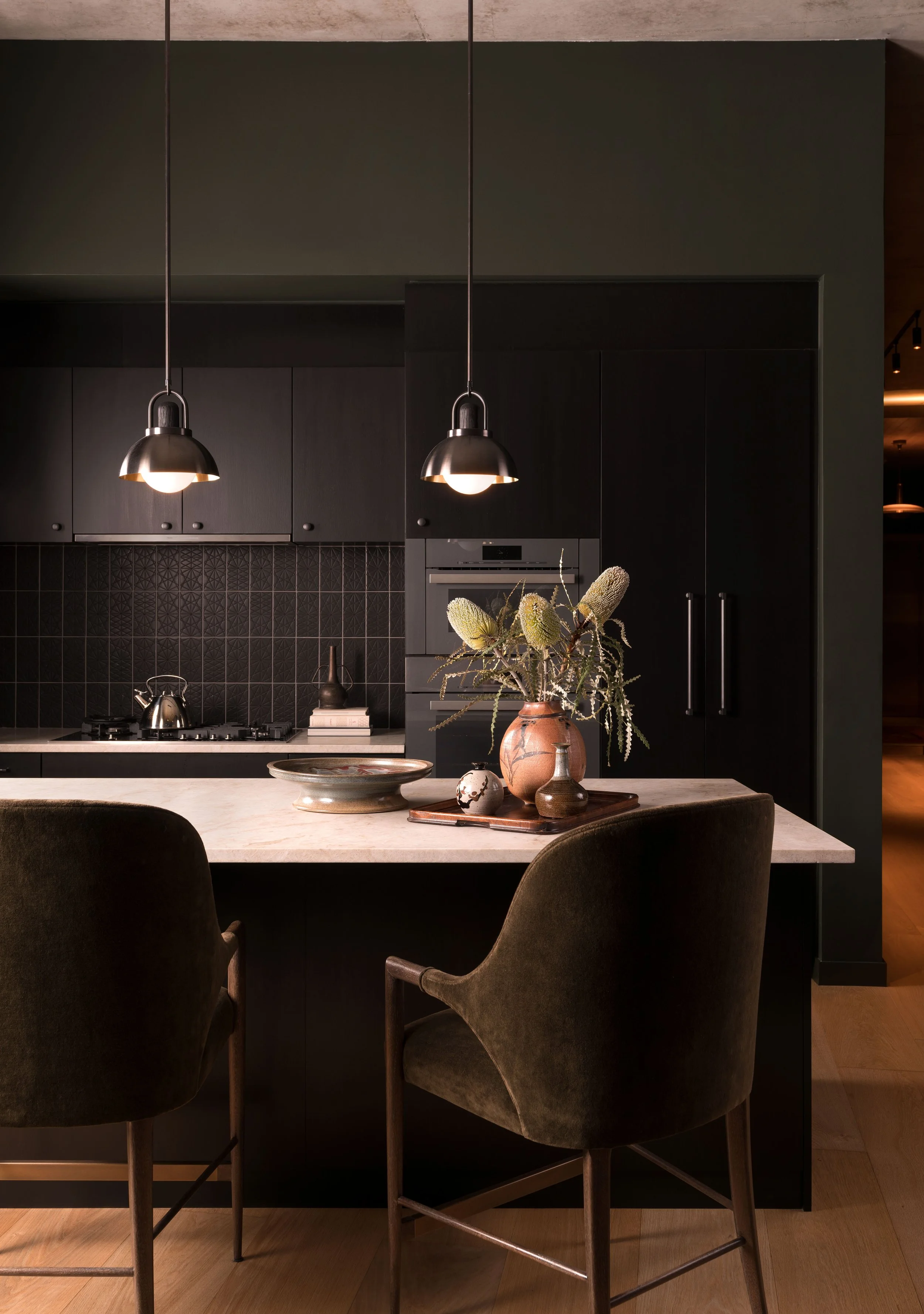  Dark, moody kitchen with black backsplash, Allied Maker pendant lights, and comfortable barstools at St. Cecilia by Cravotta Interiors. 